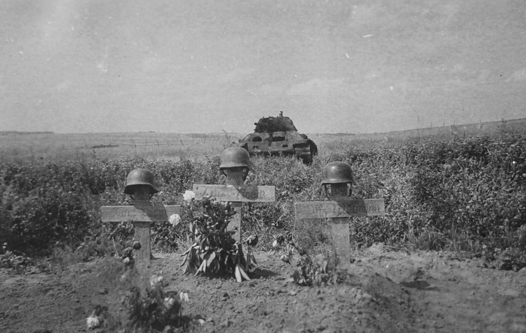 Могила немецких солдат в поле. На заднем плане стоит подбитый советский танк Т-34 образца 1940 года.
