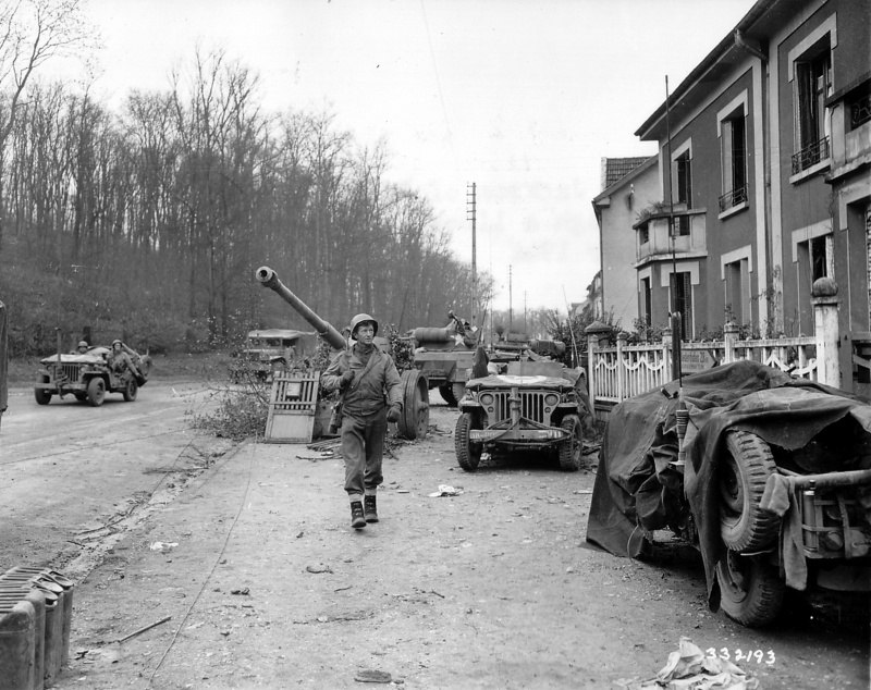 Рядовой 95-й американской пехотной дивизии Джексон идет по улице пригорода французского городка Мец мимо джипов и немецкой 88-мм пушки PaK 43.